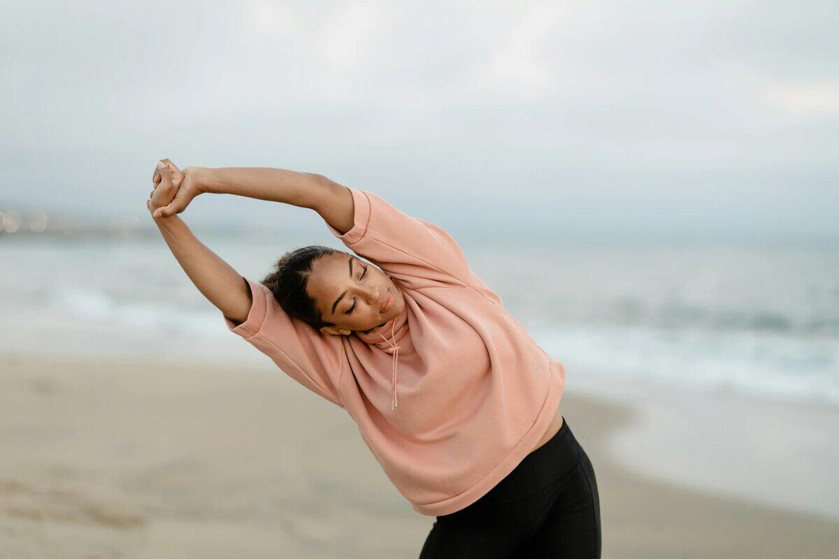 Woman in pink hoodie doing side stretch exercise on beach - health and wellness cheat sheet