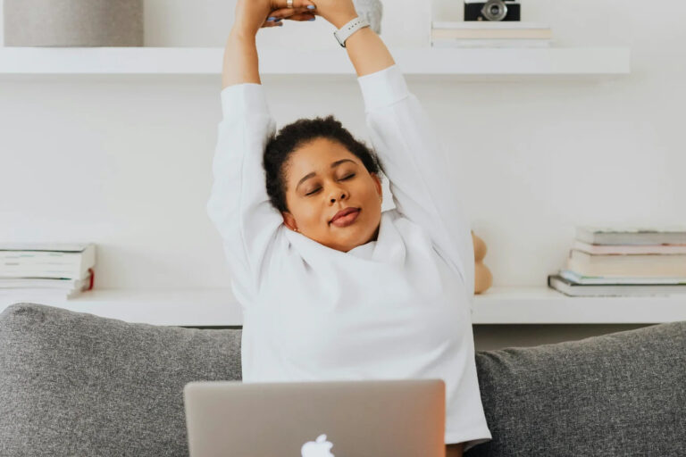 Woman in white sweater stretching her arms overhead while taking a break from working on laptop at home - afternoon energy reset